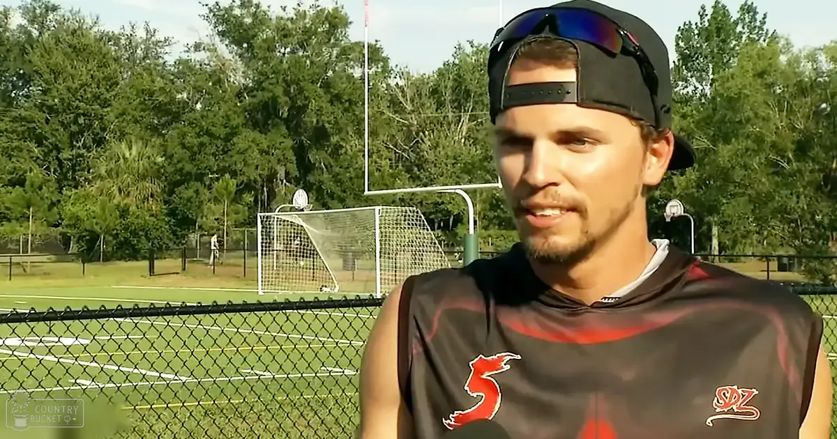 A young adult male in a sleeveless athletic jersey with the number "5" and some text, wearing a black cap and sunglasses resting on top, interviewed outdoors on a sports field with goalposts and trees in the background.