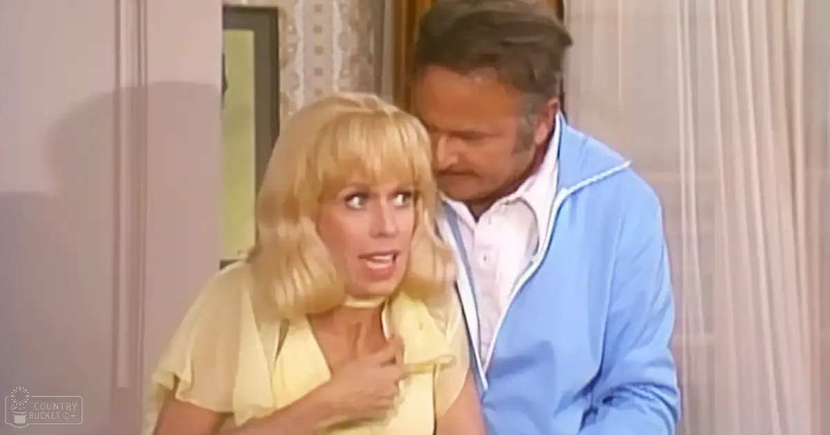 Middle-aged woman wearing a light yellow, relaxed short-sleeved blouse with ruffled collar indoors next to a middle-aged man in a light blue zip-up jacket.