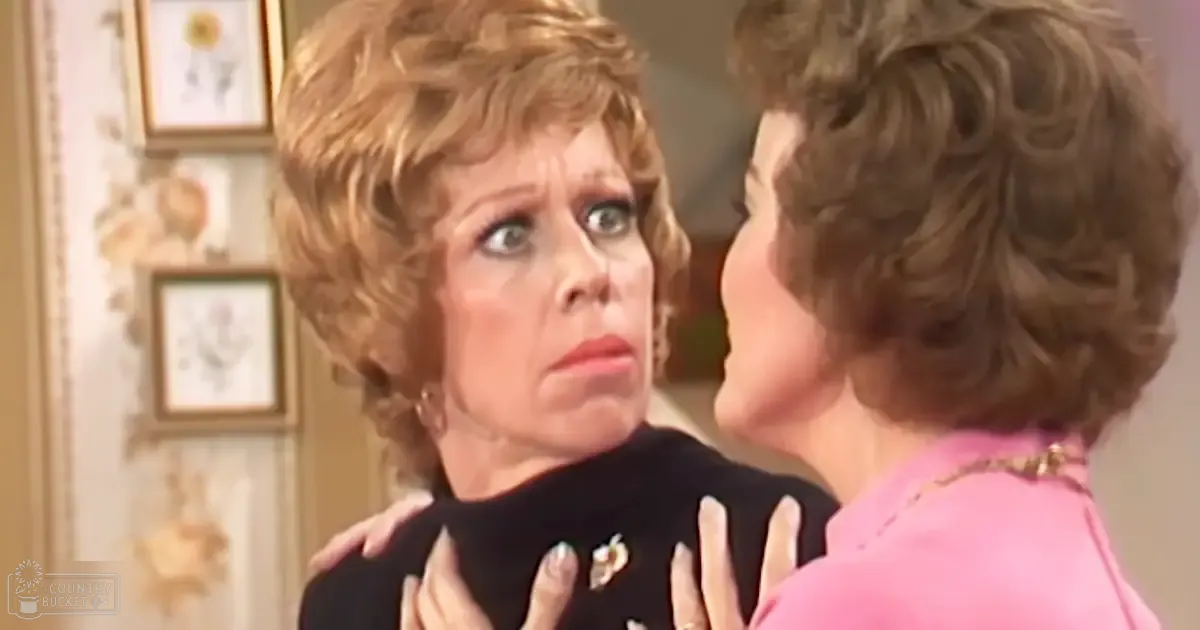 A middle-aged woman with short, curly blonde hair wearing a black top with a visible pin or brooch near the neckline stands in an indoor setting with natural daylight coming from the left side. A pink top is worn by another older adult woman in the background.