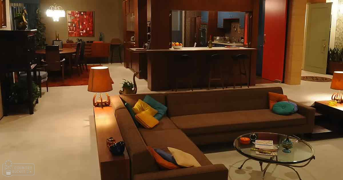 An indoor living space with a sectional sofa, side tables, lamps, glass coffee table, wooden dining area, bar counter, and piano in an apartment or house. The setting appears to be evening or night due to the dim lighting.