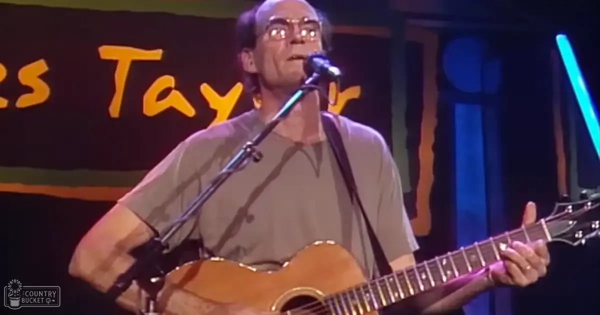 A middle-aged male singer performing at an indoor music venue with stage lighting, wearing a neutral-colored t-shirt and holding a microphone next to an acoustic guitar.