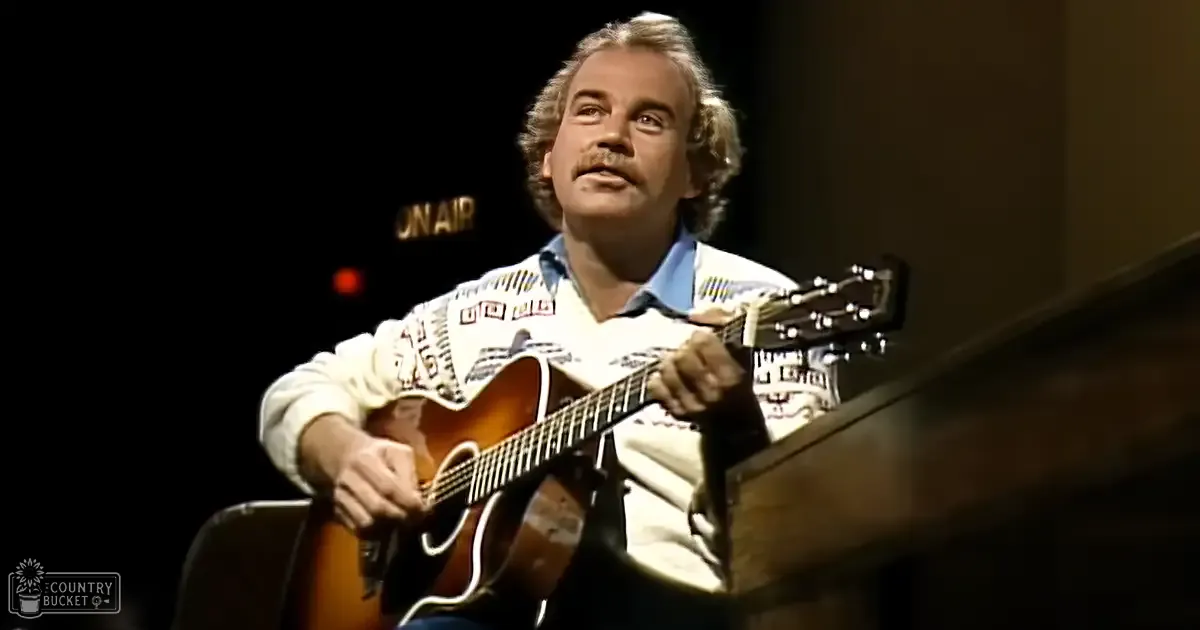 A middle-aged adult male playing a guitar in what appears to be a radio broadcast studio with an 'On Air' illuminated sign.