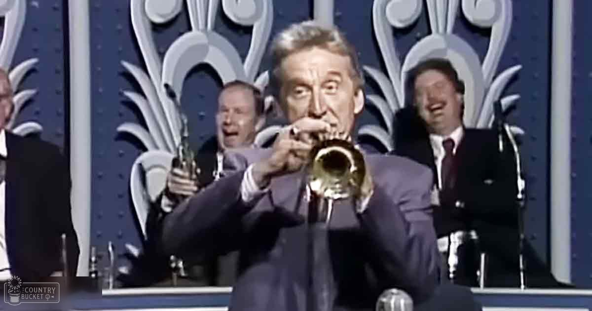 A middle-aged man playing a trumpet in what appears to be a television studio set, surrounded by other adult males dressed in dark suits and white shirts.