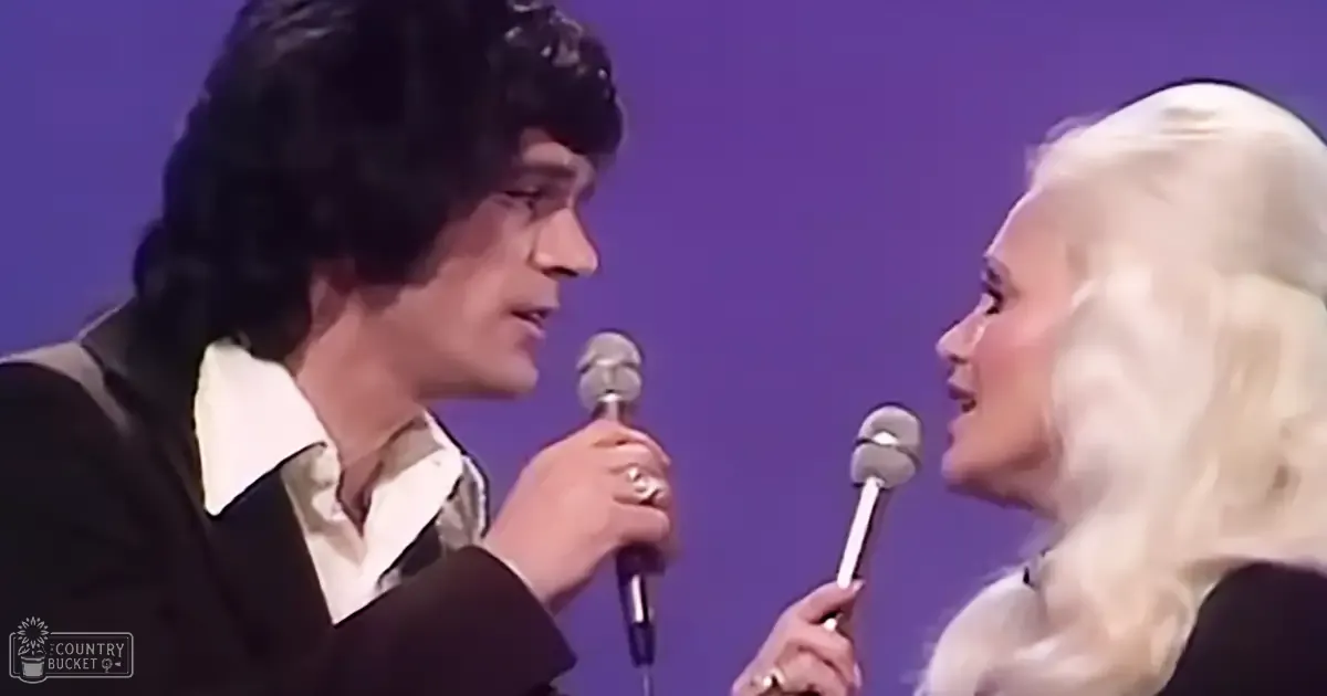 Musical performance scene with two adults, one male in a dark suit and the other female in a white dress, set against a solid purple backdrop. Microphones are held by both performers.
