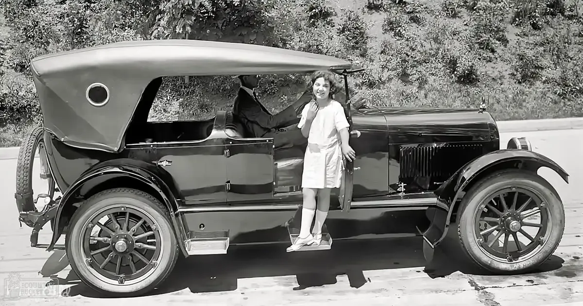 A young adult female stands next to a vintage car in a wooded rural area with midday sunlight casting shadows.