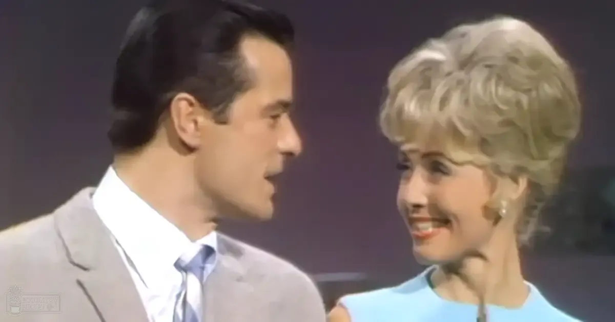 An adult male singer in a gray suit jacket, white shirt, and light blue tie is accompanied by an older female wearing a light blue dress with a high neckline indoors on a dark backdrop.