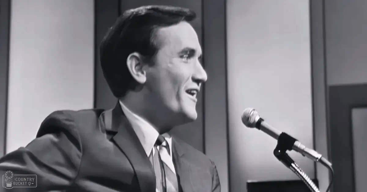 A middle-aged man wearing a dark suit jacket, white shirt, and tie, performing or speaking into a microphone in an indoor setting with simple decor.