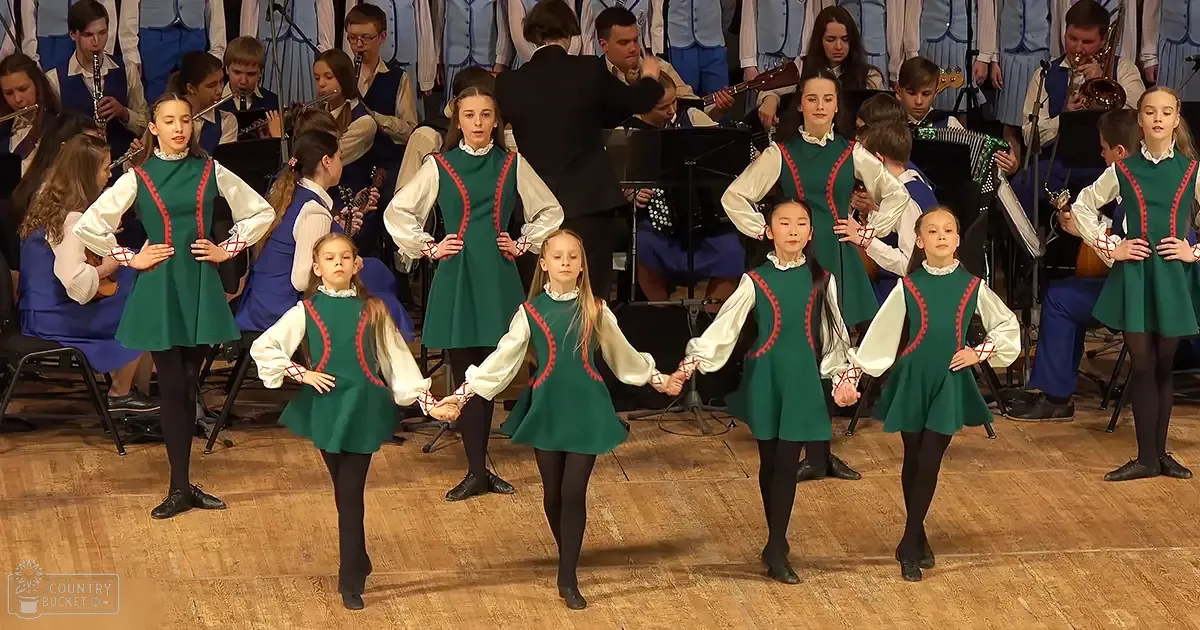 Children performing a dance in front of an orchestra at a school or community hall, children wearing green dresses with red trim and white sleeves, adults in formal attire seated behind them, bright indoor setting with musical instruments and sheet music stands.