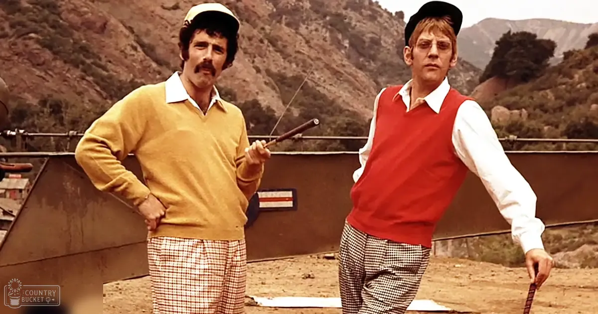 Two adult males in a rural outdoor setting, wearing mustard yellow sweaters with plaid pants and red vests with white shirts; metal structure and partially legible sign visible behind them under natural light.