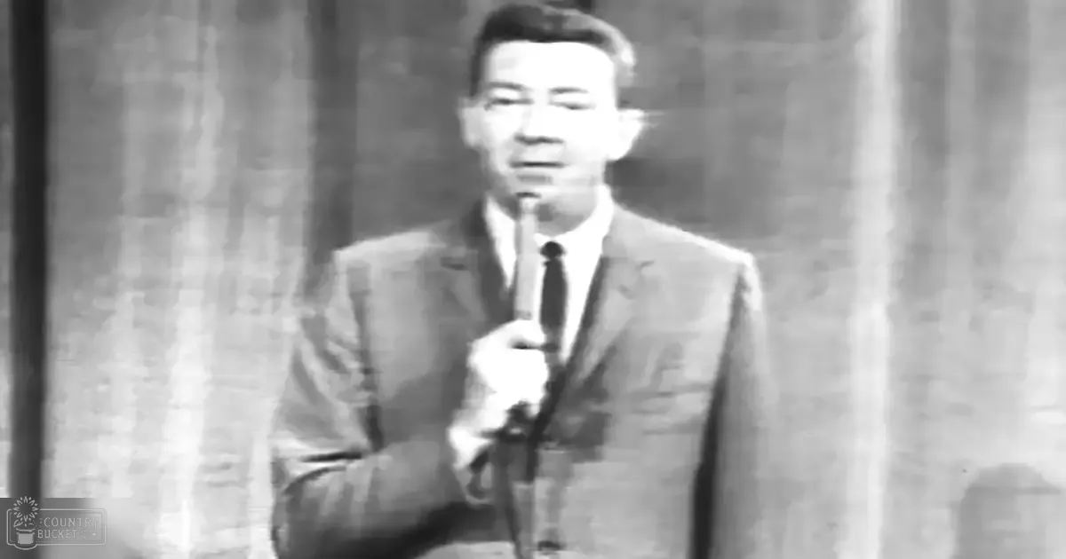 A middle-aged man wearing a well-fitted suit jacket with a notched lapel, holding a microphone in an indoor setting that suggests a stage or studio environment.