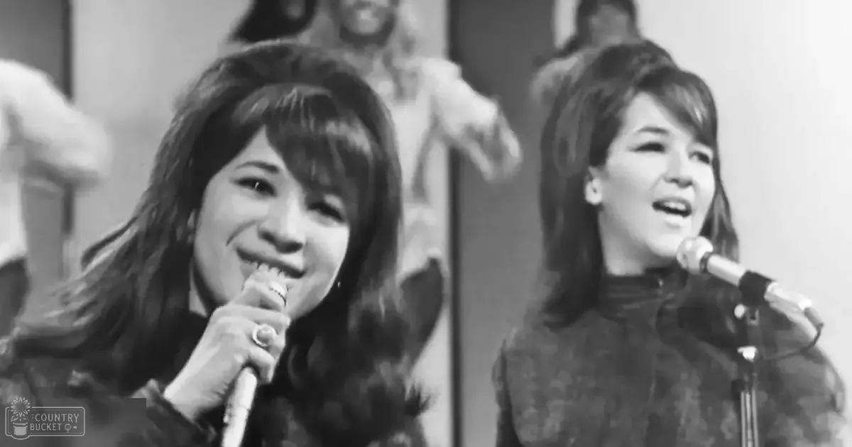 An adult female singer wearing a dark jacket with a fur-lined hood, possibly indicating winter attire, stands beside another adult female in a dark dress or top with a high neckline and long sleeves. The setting appears to be an indoor performance venue with stage lighting.