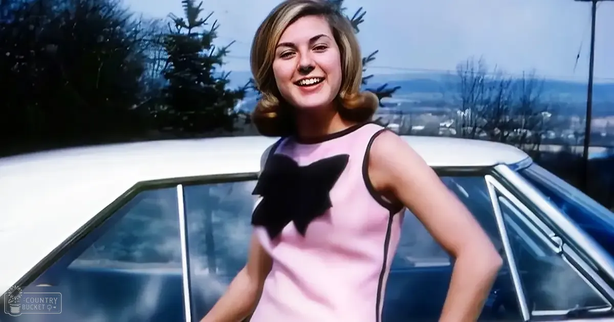 A young adult female in a sleeveless black bow dress with a light fabric stands outdoors, surrounded by trees and distant buildings under partly cloudy skies.
