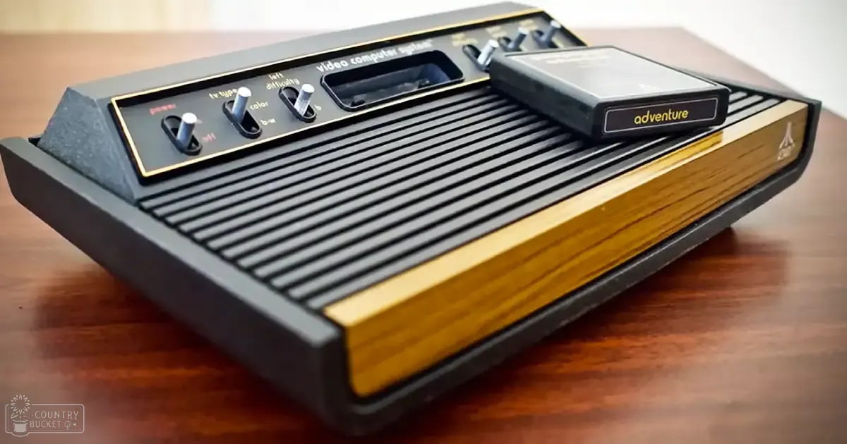 Indoor scene with vintage Atari 2600 video game console, wooden surface, small black cartridge labeled 'adventure', two control sticks visible on top of the console.