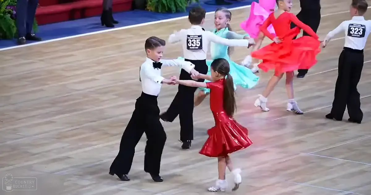 Children participating in a dance competition at an indoor venue with wooden floors, red seating, and greenery in the background.