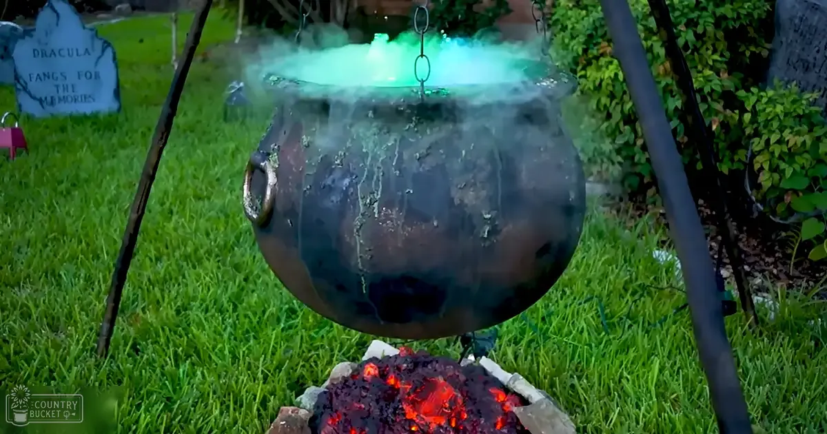 A Halloween-themed outdoor scene with a large black cauldron with steam rising, glowing embers in a fire pit below, and a tombstone reading 'Dracula Fangs For The Memories' amidst greenery.