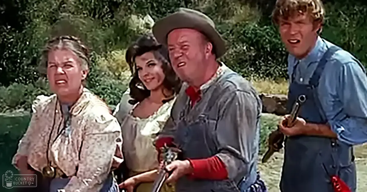 A group scene from a Western movie set outdoors near a riverbank, with lush greenery suggesting spring or summer and natural daylight illuminating the group.