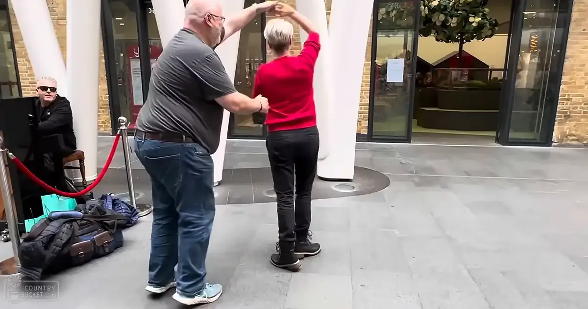 A middle-aged man wearing a dark gray t-shirt and blue jeans dances with an adult female in red top and black pants, set against urban backdrop with red velvet rope barrier.