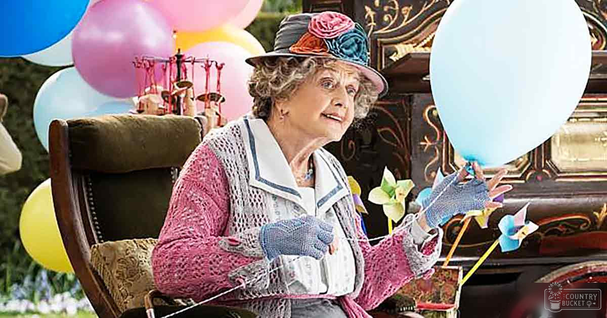 An older adult female wearing a pink knitted sweater vest over a grayish-blue shirt, accessorized with fingerless gloves and an ornate floral hat, in a whimsical indoor setting decorated for a celebration with balloons.