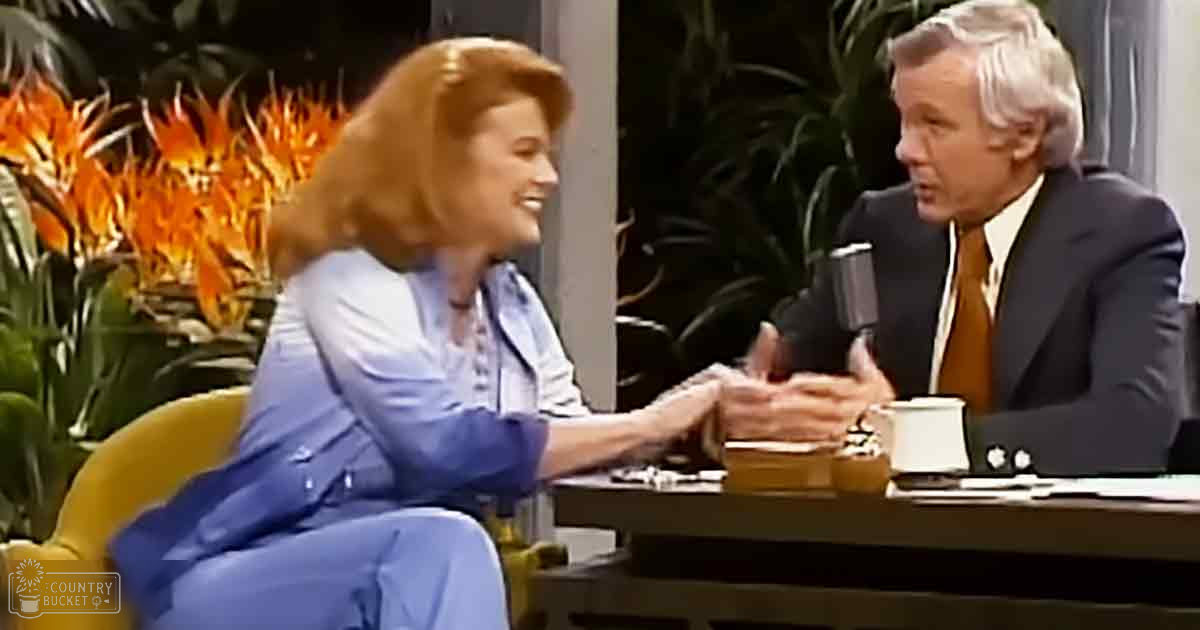Two adults seated at a table, likely an indoor studio setting with greenery and orange flowers, suggesting a talk show or interview environment.