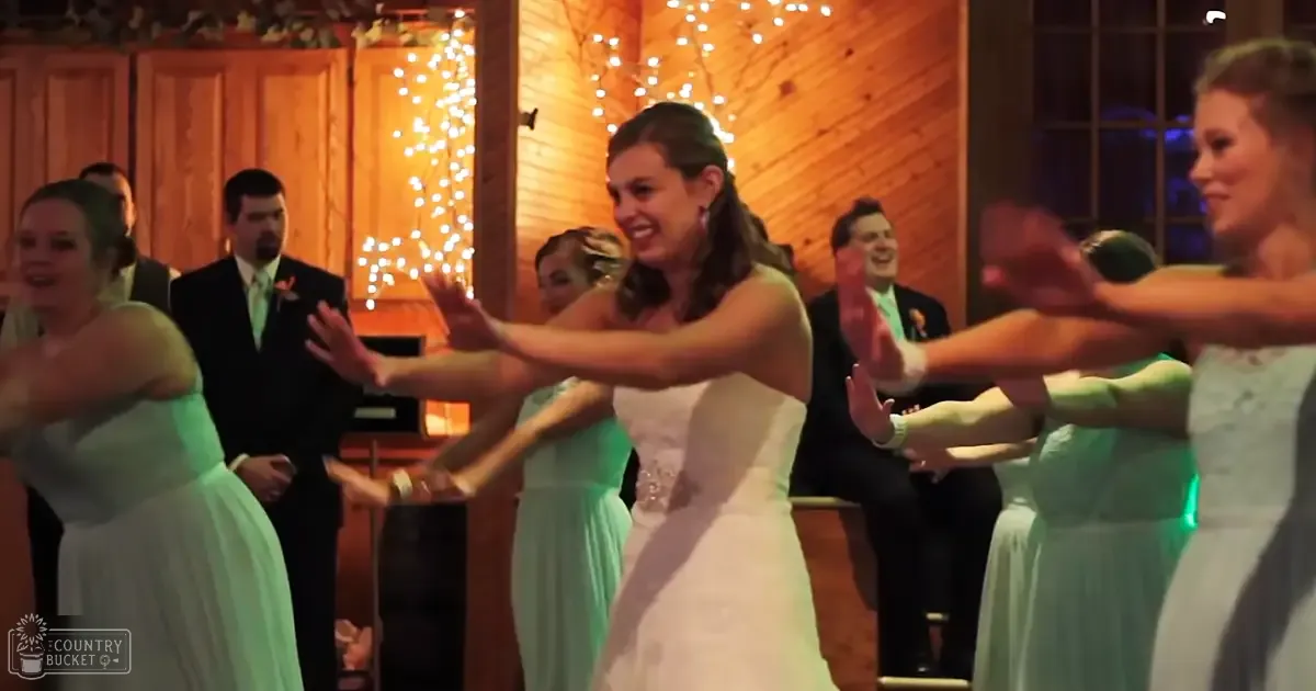 A young bride dancing with bridesmaids in a rustic indoor wedding reception, surrounded by wooden walls and hanging string lights.