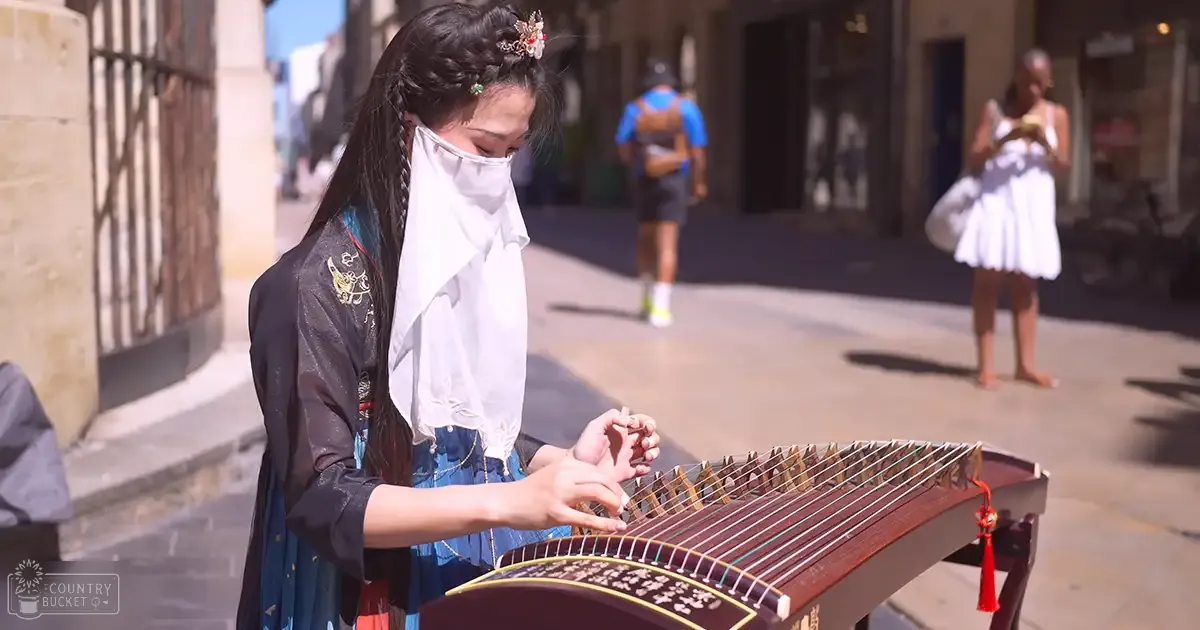 A young adult female plays a guzheng on an urban street with another adult male and a younger or older female in the background, wearing traditional Chinese attire. The scene is set during summer with clear blue skies.