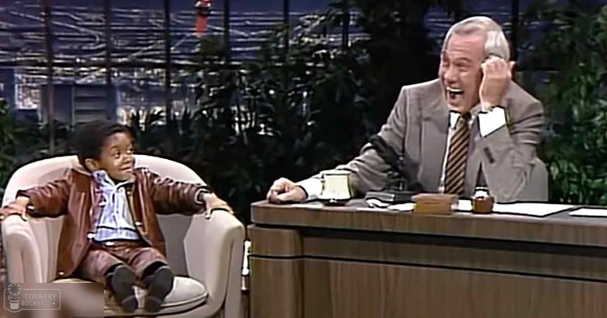 A toddler wearing a brown leather jacket over a blue and white checkered shirt sits on a white armchair against an urban night backdrop with green plants. An older adult male in a light gray suit sits behind a wooden desk, lit by artificial studio lighting.