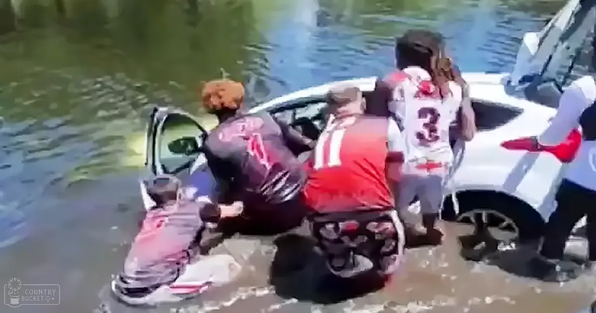 A flooded area with a white car partially submerged in water; several adults wearing football jerseys are present, indicating an outdoor daytime setting near a river or canal.