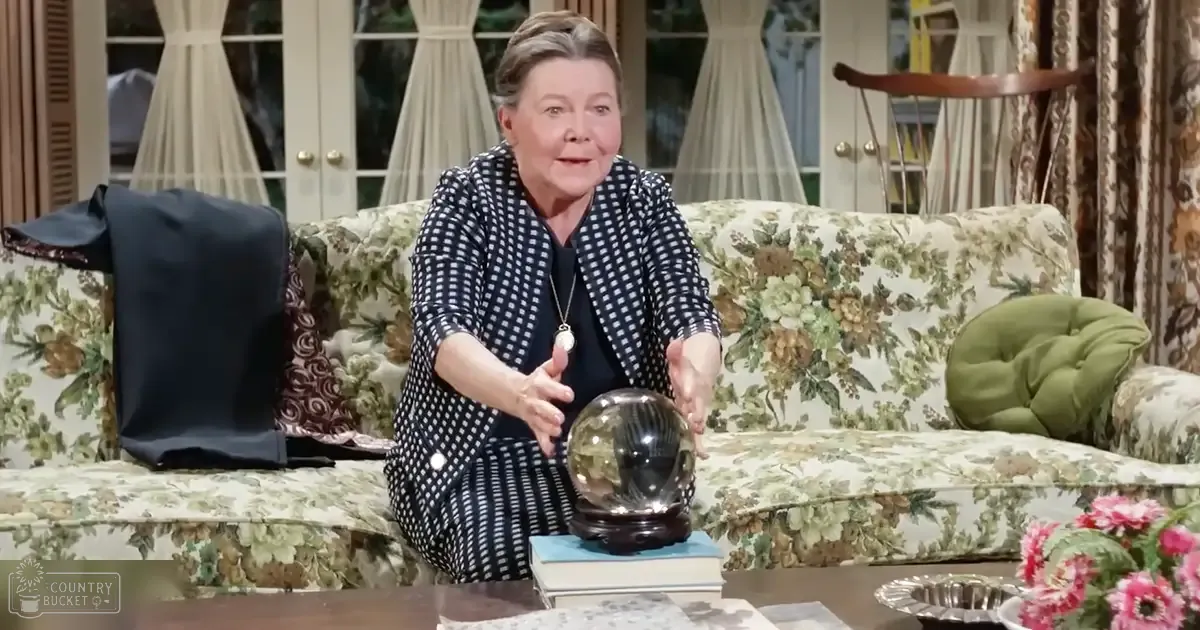 Older adult female in a patterned dress with a blazer over it, sitting on a floral-patterned couch in a living room with spring or summer decor and natural daylight.