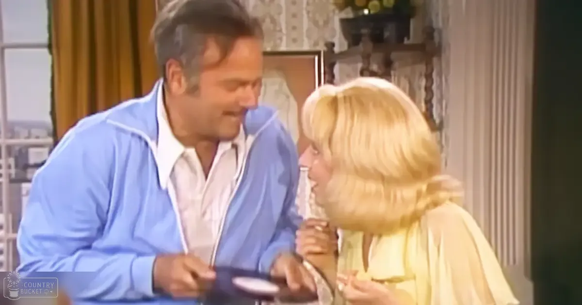 An adult male singer in a light blue zip-up cardigan over a white collared shirt with an adult female wearing a pale yellow dress or robe stands indoors near ornate furniture and patterned wallpaper, holding a glass of wine; natural lighting suggests daytime.