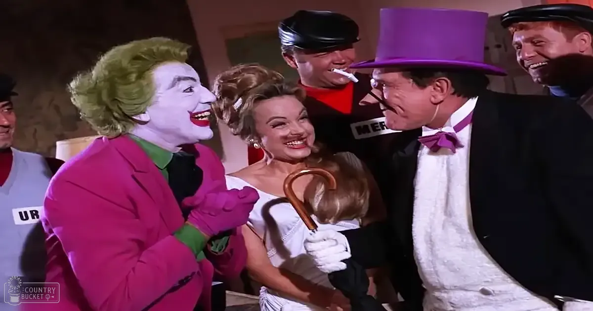An adult male character in a bright pink suit jacket and pants, surrounded by other individuals dressed in period costumes indoors, possibly on a theatrical stage.
