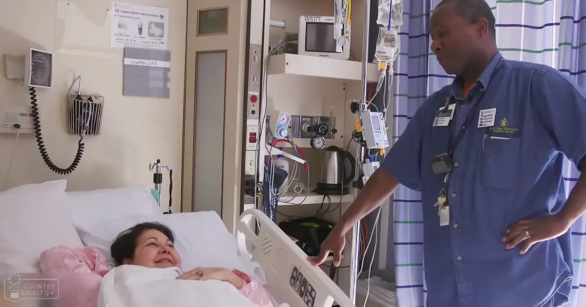 A middle-aged to older adult female patient lying in a hospital bed with white sheets and pink blankets, wearing a simple cotton hospital gown; next to her stands a middle-aged male medical professional in a blue button-up shirt with 'Together Caring' badge.