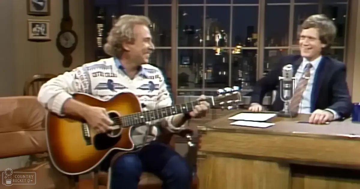 A middle-aged man wearing a white sweater with blue patterns, playing guitar in a talk show setting.