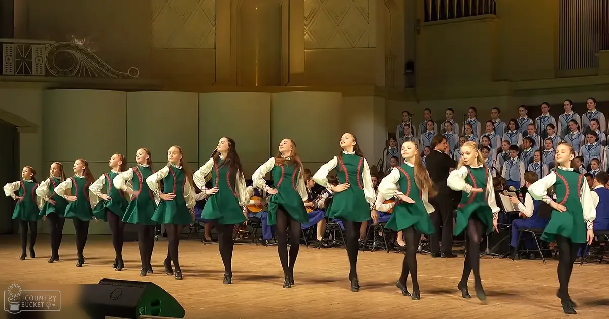 Young female dancers performing on stage with green dresses, blue uniforms, and a large organ in the background under bright stage lighting.