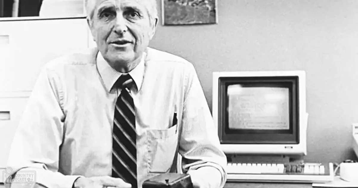 An older adult male in a professional office setting wearing a button-down shirt and striped tie with an old computer monitor behind him.
