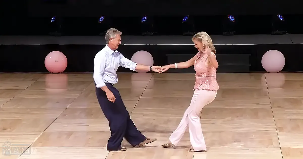 A middle-aged to older adult couple dancing indoors, with a man in a white dress shirt and dark blue trousers, and a woman in a pink lace top and light pink pants. The setting appears to be an event or performance space with wooden floors and stage lighting.