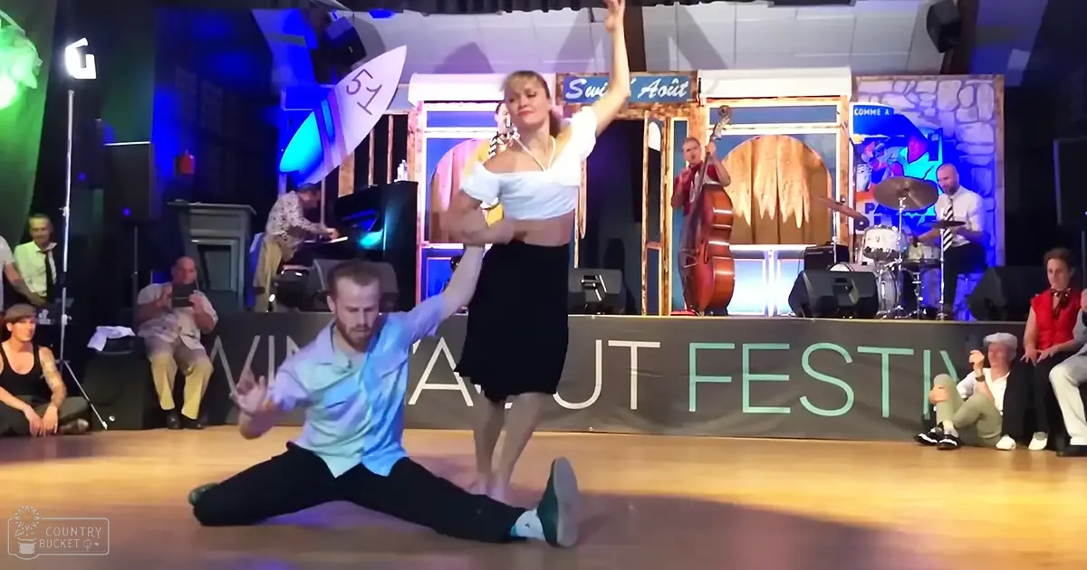 A dance duo performing at the Swing About Festival in an indoor dance hall with wooden flooring, a stage area, and audience seating.