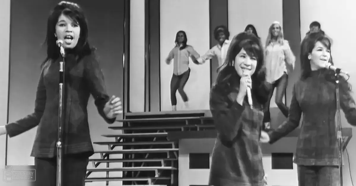 A music video or television show performance scene set in a modern studio environment with geometric patterns on the backdrop, with young adult females wearing dark-colored jackets and light-colored blouses under artificial lighting.