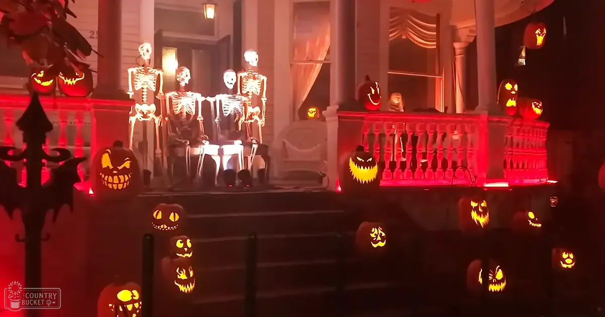 Halloween-themed porch scene with illuminated jack-o'-lanterns, skeleton figures, red lighting, seasonal decorations, residential neighborhood at night or early evening.