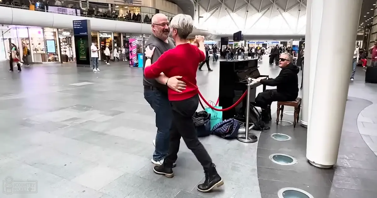 A middle-aged couple dances in a public indoor space with shopping bags visible near a seated older adult pianist, surrounded by pillars and high ceilings.