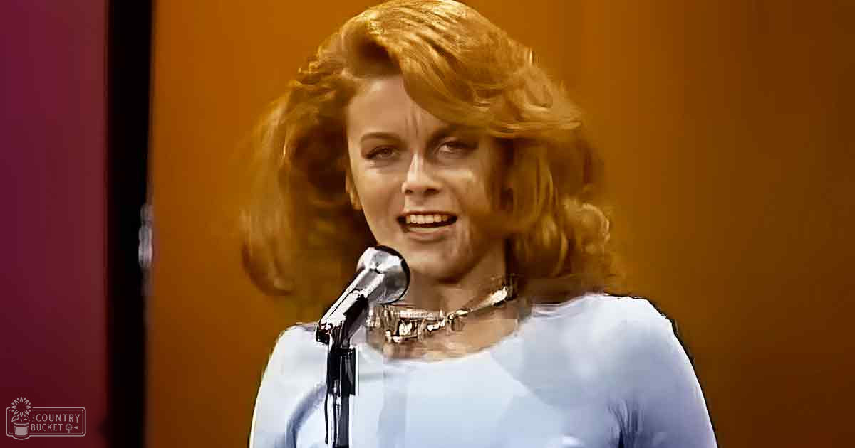 A young adult female wearing a light blue, relaxed-fit long-sleeved blouse with a round neckline, accessorized with an intricate choker-style necklace, stands in front of a microphone and door frame against a reddish-brown wall under warm lighting.