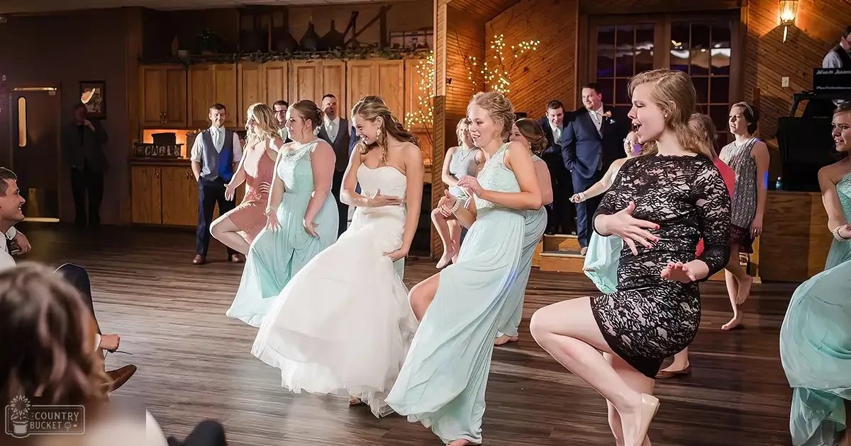 A rustic indoor wedding reception scene with wooden dance floors, decorative garlands on cabinets, string lights for evening ambiance, light blue bridesmaids' dresses, white bride's gown, and a variety of formal attire among guests.