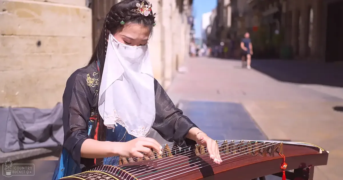 A young adult female playing a guzheng in an urban street setting with stone buildings and people walking by.