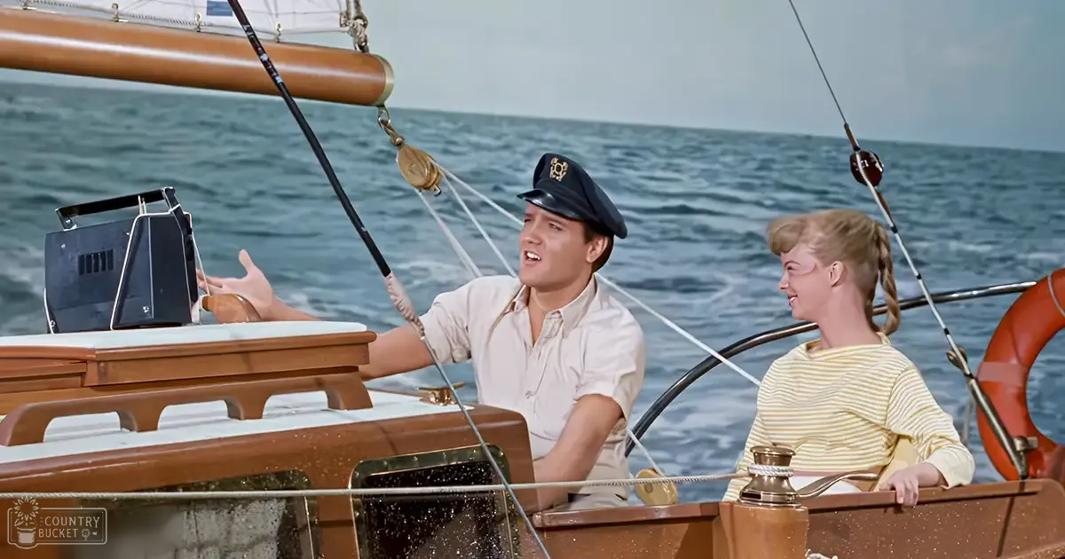 A young adult male wearing a light-colored shirt and captain's hat on a wooden sailboat amidst gentle waves under an overcast sky.