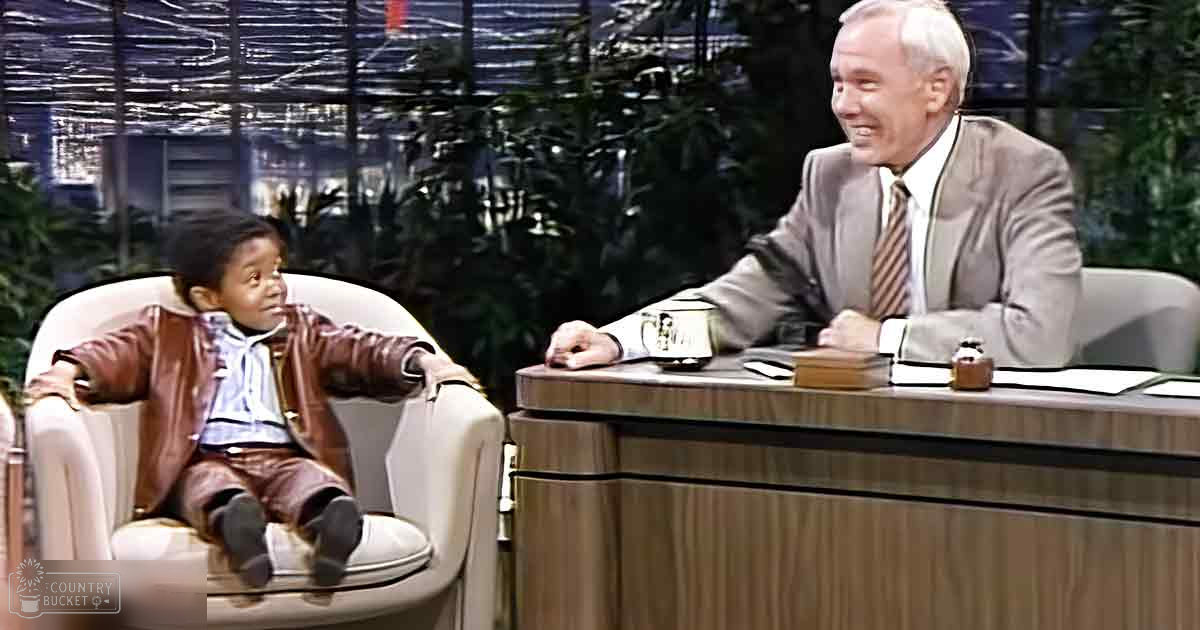 A young toddler in a brown leather jacket and white hoodie sits beside an older adult male dressed in a light gray suit, against a backdrop of indoor greenery with city views.