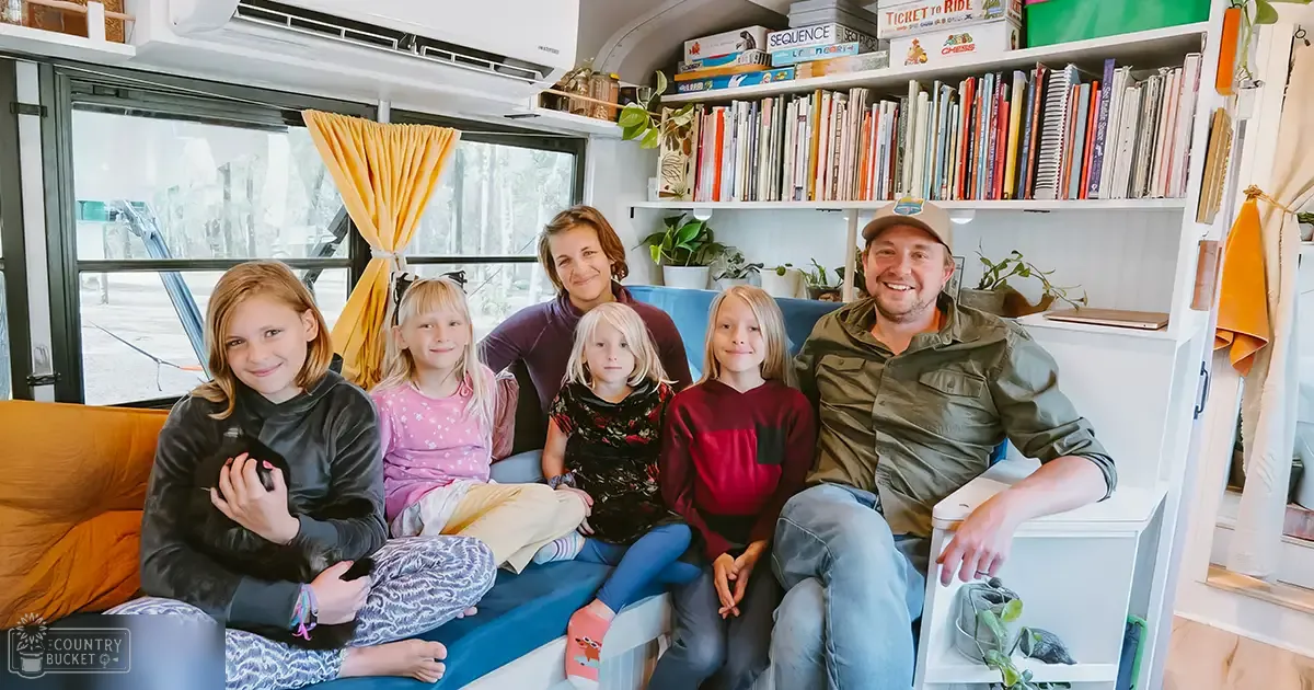 A family of six, including children and an adult, inside a bright RV interior with large windows showing overcast daylight. A small kitten or puppy is held by one child.