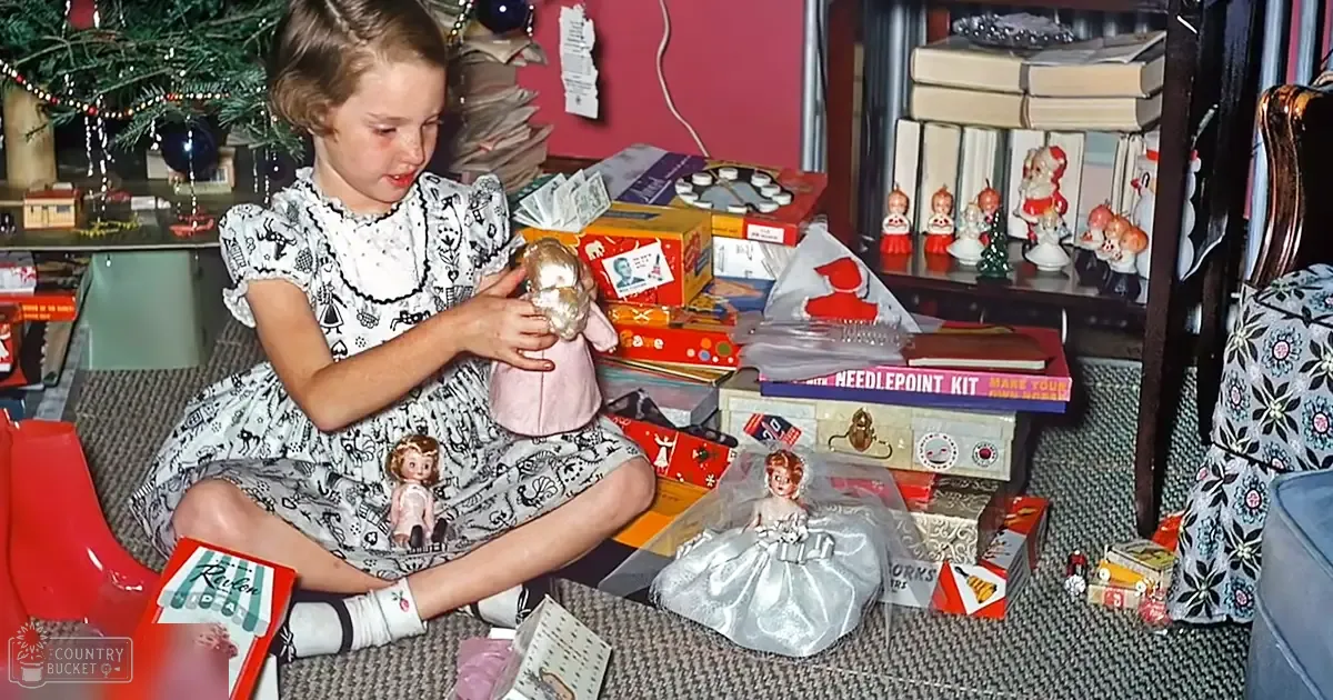 A young girl in a patterned dress with black and white cat print, seated indoors surrounded by holiday decorations including a Christmas tree and wrapped gifts.