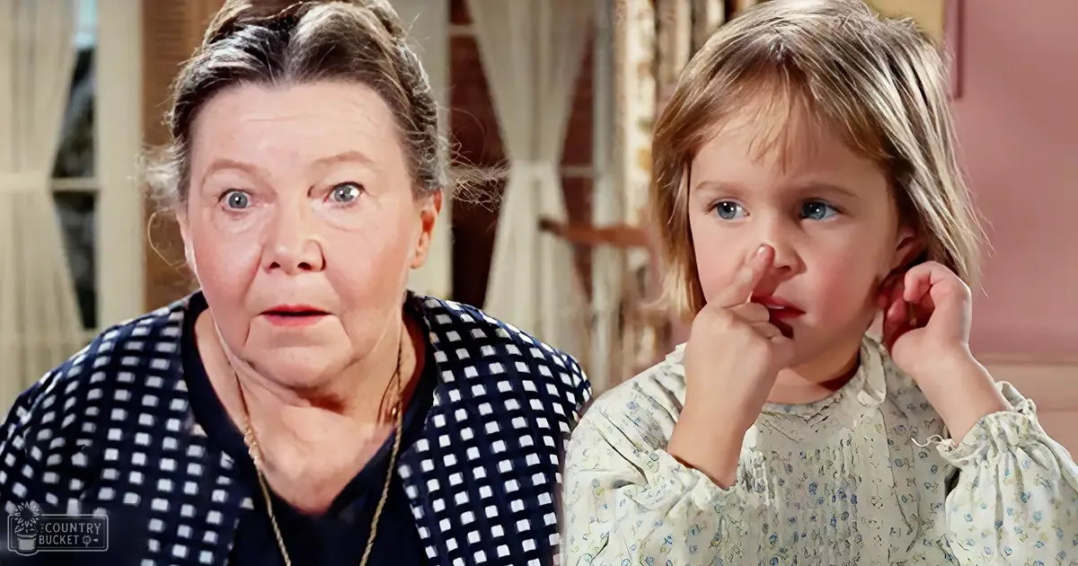 An older adult woman wearing a dark blue garment with white polka dots and a light-colored cardigan indoors with a toddler holding her nose and touching her ear, suggesting a casual home environment during the daytime.