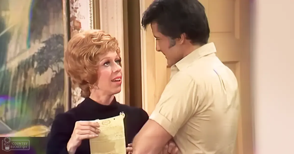 Middle-aged woman in a black zip-up turtleneck sweater indoors, holding a yellow envelope with text partially visible; light beige polo shirt on adult male in the background.