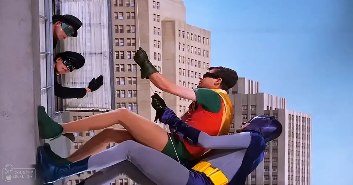 A superhero in action, wearing a vibrant green and orange costume with gray pants and yellow belt, facing off against an opponent dressed in dark clothing. The scene is set in an urban environment during the day.