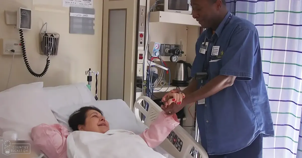 Middle-aged to older adult female in a hospital room, relaxed and smiling; adult male staff member in blue denim shirt supporting her hand.